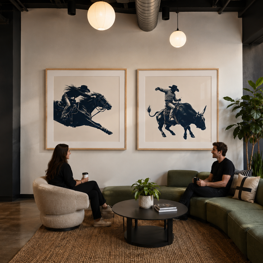 Two framed black-and-white art prints hang on a white wall in a modern lounge setting, with two people seated on a green sectional sofa and a round coffee table in front. The scene includes plants, pendant lights, shelving, and an industrial ceiling with exposed ductwork.