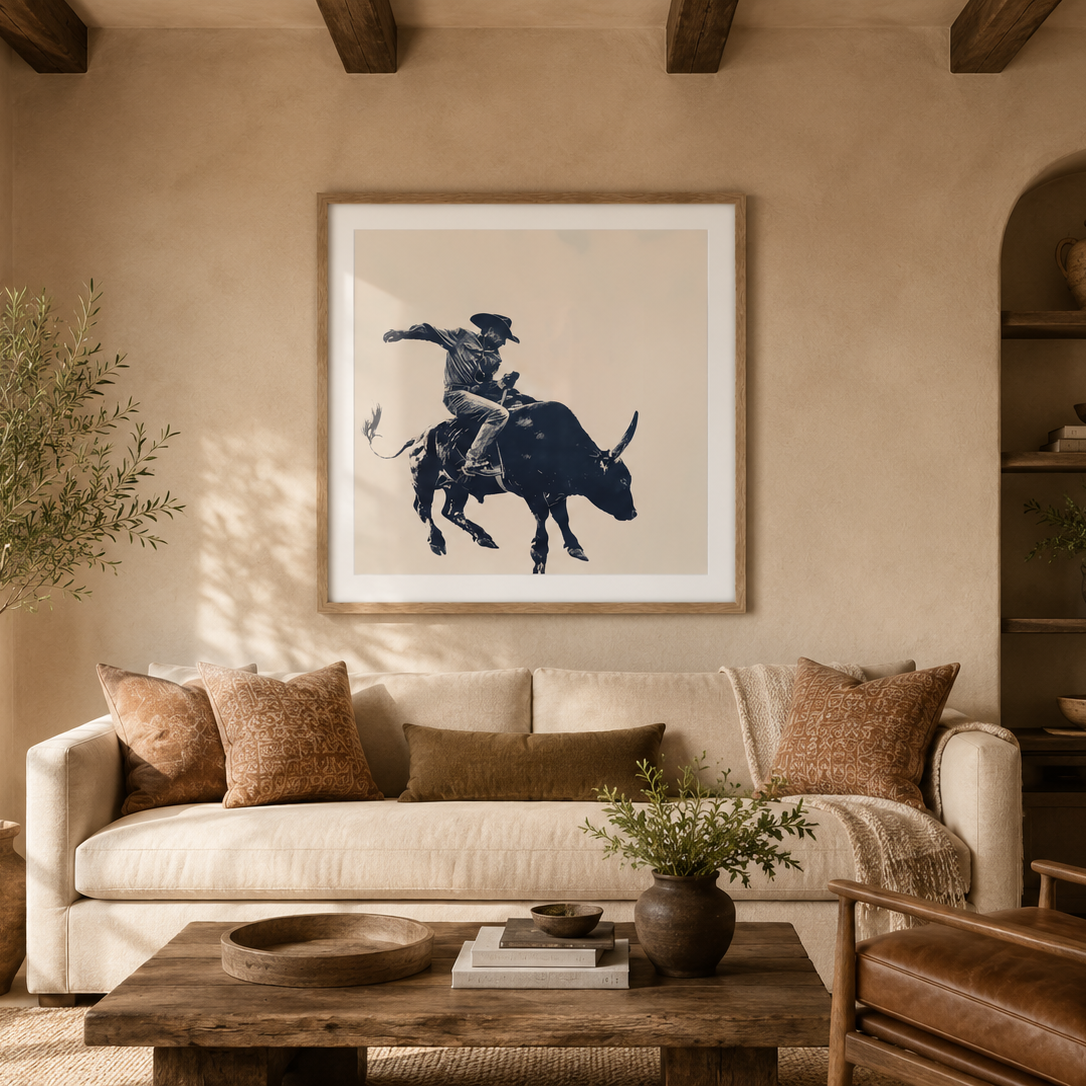 A framed art print featuring a black ink illustration of a cowboy riding a bucking bull, displayed on a warm beige wall in a rustic, sunlit living room with a cream sofa, wooden coffee table, and leather chair.